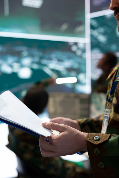 Militaire en uniforme camouflé tenant un dossier dans un centre de commandement, avec des écrans de contrôle et d'autres personnels en arrière-plan
