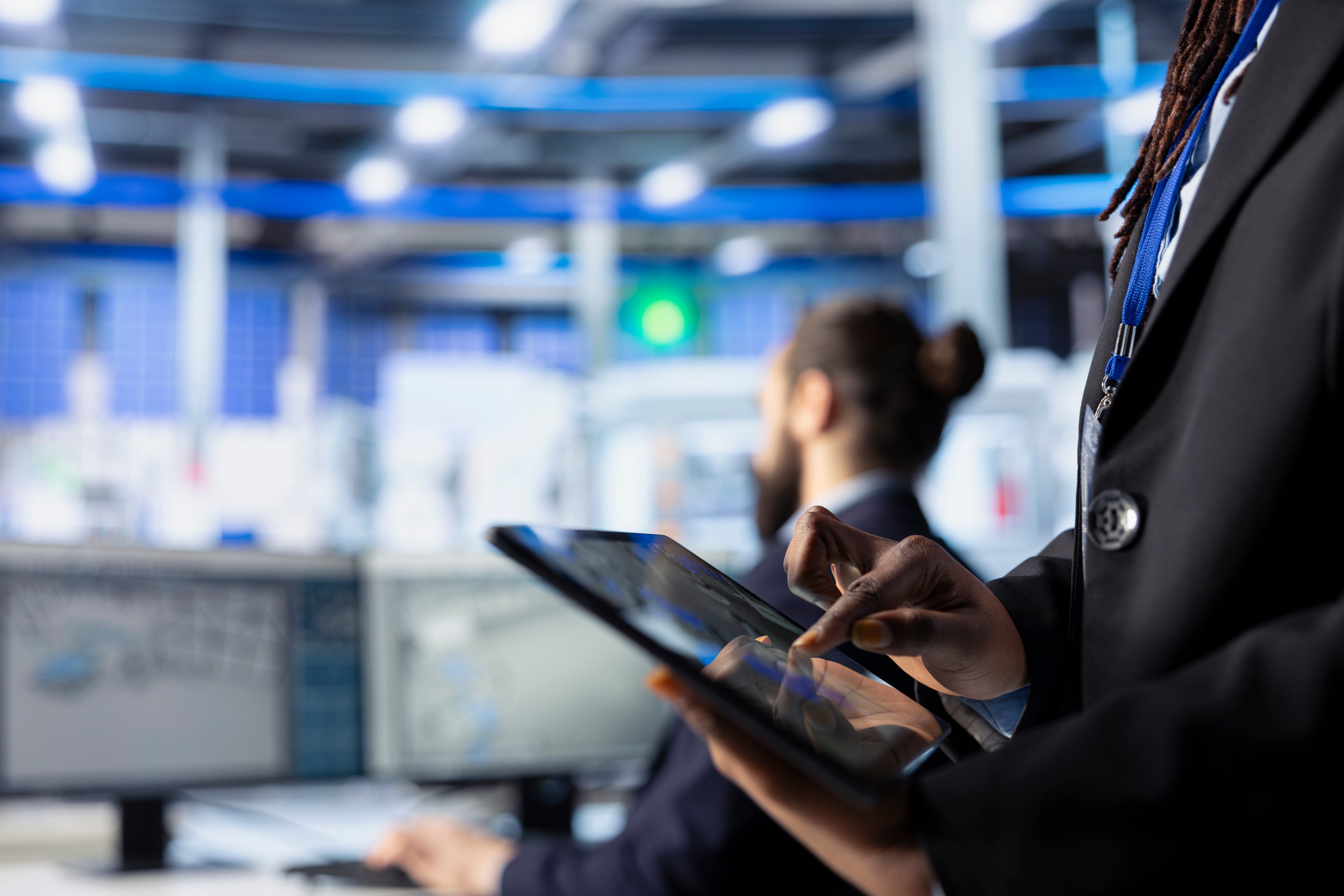 Professionnel du secteur de la défense utilisant une tablette tactile dans un centre de contrôle technologique, avec des écrans de surveillance en arrière-plan.